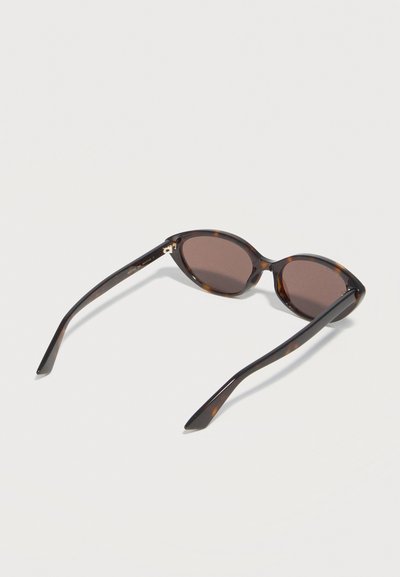 Οβάλ γυαλιά ηλίου με σχέδιο χελώνας και σκούρους καφέ φακούς, ανοιχτά σε ελαφρύ υπόβαθρο, με κλίση για να φαίνεται το πλαίσιο και τα μπράτσα.