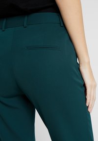 Tealfärgade byxor i ett slätt tyg med smal passform. Har en dekorativ bakficka och en platt midja.