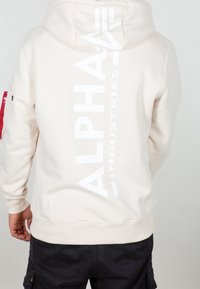 Béžová mikina s velkým bílým textem "Alpha Industries" na zádech, model otočený zády, oblečený v černých cargo kalhotách v jednoduchém studiovém prostředí.
