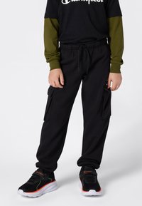Zwarte cargo broek met zijzakken, elastische tailleband. Draagt een zwart shirt en een groene lange mouwlaag. Zwarte en rode sneakers.