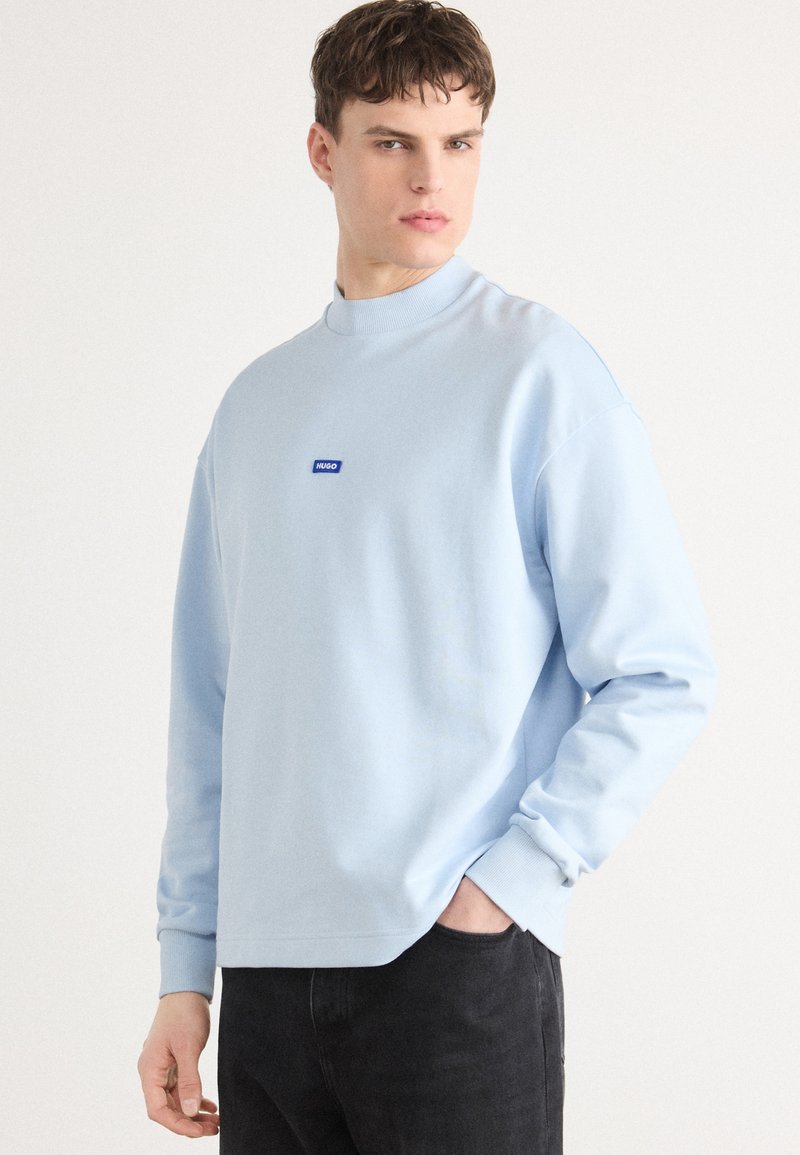 Felpa azzurra con collo alto e maniche lunghe, caratterizzata da un piccolo logo blu "HUGO" sul petto, abbinata a jeans neri.