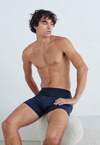 Mørkeblå boxershorts laget av myk, elastisk stoff, med en bred linning og en tettsittende design, vist på en sittende mannelig modell.