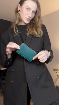 Porta-cartões verde-azulado com fecho de zíper, material em couro, formato retangular e detalhe de logótipo subtil. Modelo veste um casaco preto.