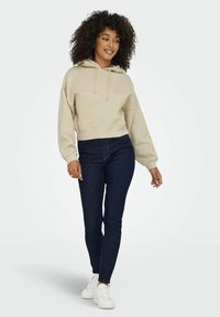 Beige verkürzter Kapuzenpullover mit Kordelzügen und überschnittenen Schultern, kombiniert mit dunkelblauen Skinny-Jeans und weißen Sneakers. Glatte Stofftextur.