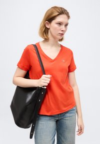 Νεαρή γυναίκα με πορτοκαλί μπλουζάκι V-λαιμόκοψη και ανοιχτόχρωμα τζιν, κρατώντας μια μεγάλη μαύρη τσάντα ώμου με λουρί πάνω στον ώμο της.