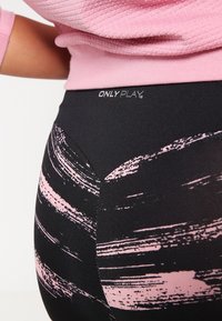 Zbliżenie na legginsy w czarno-różowy abstrakcyjny wzór z logo "ONLY PLAY" w talii, noszone z teksturowanym różowym topem.