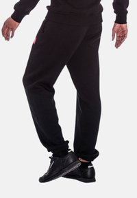 Schwarze Jogginghose aus softem Material, mit Seitentaschen und elastischen Bündchen. Getragen mit schwarzen Sportschuhen. Einfaches Design.