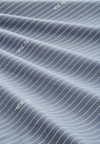 Baumwollstoff mit einem hellblauen Hintergrund und dünnen, weißen horizontalen Streifen. Hat eine glatte Textur und dezente Markenkennzeichnung in der Ecke.