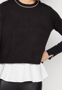 Suéter negro de punto con una capa de peplum blanca en la parte inferior. Acentos plateados en el escote y los bordes de las mangas. Dobladillo de canalé texturizado.