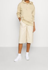 Sudadera beige con capucha ajustable y bolsillo tipo canguro, combinada con pantalones cortos anchos en tono beige claro y zapatillas blancas. Textura suave, diseño casual.