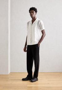 Cămașă albă cu mâneci scurte, din tricot deschis, cu modele texturate, asortată cu pantaloni negri cu croială lejeră și pantofi negri, pe un fundal de pardoseală din lemn.