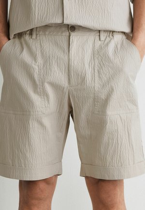 Beige strukturierte knielange Shorts mit Seiten- und Vordertaschen, getragen von einer Person mit den Händen in den Taschen, kombiniert mit einem passenden beigen Oberteil.