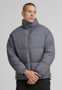 Dunkelgraue Pufferjacke mit hohem Kragen, Reißverschluss und elastischen Bündchen. Weicher Stoff mit gestepptem Muster zur Isolierung.