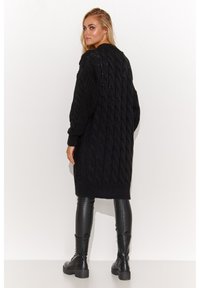 Czarny, oversize sweter w splot warkoczowy z długimi rękawami, teksturowanym wzorem i ściągaczem na dole, noszony na czarnych, dopasowanych leginsach i botkach.