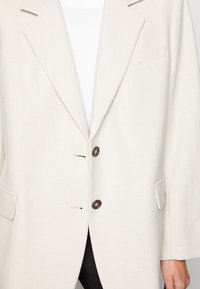 Blazer bianco in lana con rever a contrasto, botttoni doppi e tasche laterali. Presenta una finitura tessurizzata e uno stile frontale aperto.