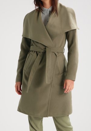 Woman wearing an olive green belted coat with wide lapels over matching pants and a light gray shirt.