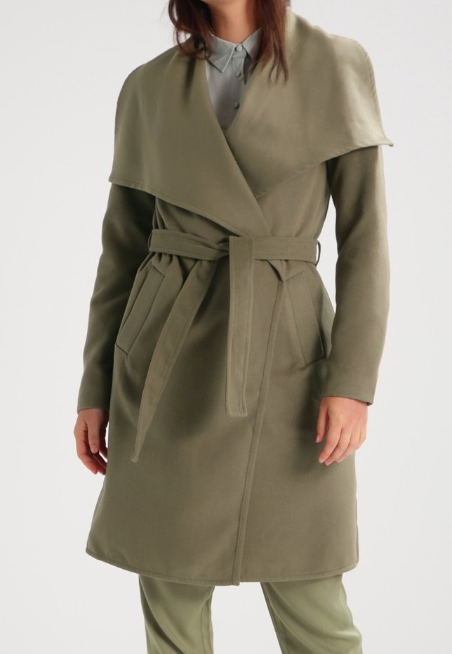 Femme portant un manteau ceinturé vert olive à larges revers sur un pantalon assorti et une chemise gris clair.