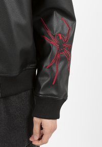 Giacca in pelle nera con finitura texturizzata e polsini a costine. Presenta un vistoso ricamo rosso a forma di ragno sulla manica sinistra.