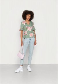 Top floral verde com mangas curtas com babados, combinado com jeans azul claro e ténis brancos. Segurando uma pequena carteira rosa.