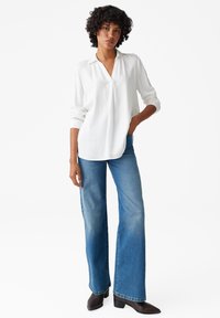 Weiße Bluse mit langen Ärmeln, Kragen und V-Ausschnitt, kombiniert mit blauen Schlagjeans und schwarzen Stiefeletten. Glatte Textur und lockere Passform.