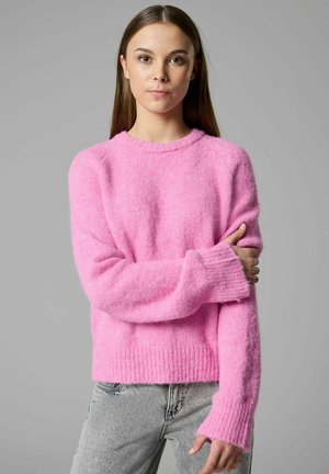 Fiatal nő egyenes barna hajjal, rózsaszín bolyhos pulóvert és világosszürke farmert visel, karját keresztbe téve áll szürke háttér előtt.