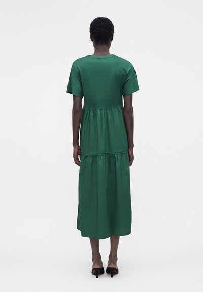 Άτομο που στέκεται με την πλάτη, φοράει πράσινο midi φόρεμα με κοντά μανίκια και μαύρα παπούτσια με τακούνι σε λευκό φόντο.