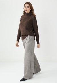 Brauner, gestrickter Rollkragenpullover mit gerippten Bündchen, kombiniert mit weit geschnittenen, hellgrauen Hosen und schwarzen spitz zulaufenden Schuhen.