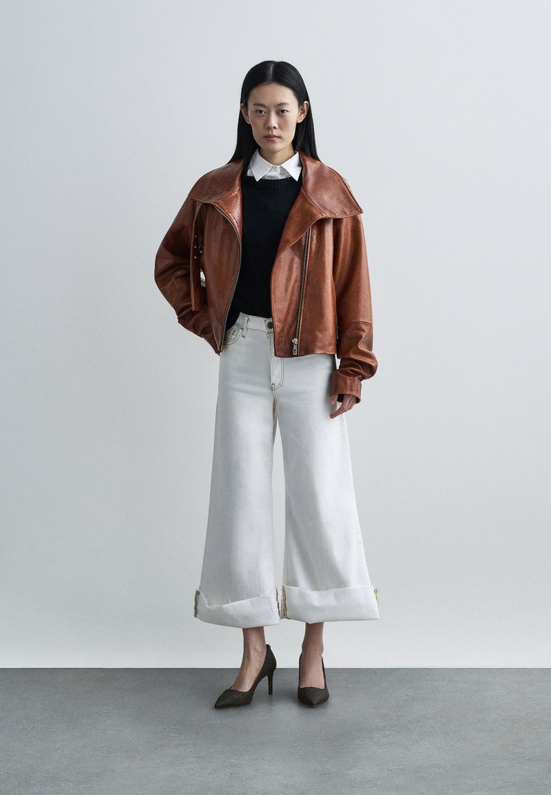 Veste courte en cuir marron avec col surdimensionné, pull noir, jean blanc large avec ourlets effilochés, et escarpins noirs pointus.