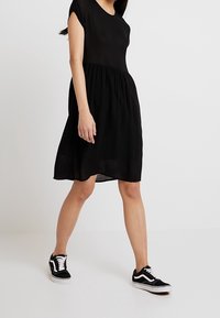 Schwarzes kurzärmeliges Kleid mit taillierter Taille und fließendem Rock; kombiniert mit schwarz-weißen Sneakern. Glatte Stoffoberfläche.