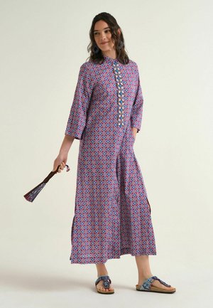 Donna che indossa un abito lungo con motivi blu, rossi e bianchi, tiene un ventaglio coordinato e indossa sandali con perline.