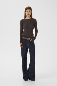 Young woman stands facing forward wearing a brown long-sleeve top, dark blue wide-leg jeans with a brown belt, and black pointed shoes.