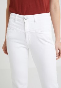 Mujer vestida con jeans blancos de tiro alto y una blusa de color óxido metida por dentro, de pie con los brazos relajados a los lados.