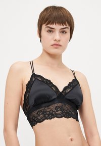 Svart bralette laget av satengstoff med blondebesetning langs koppene og kanten, justerbare tynne stropper, og blomsterblonde mønster ved bunnen.
