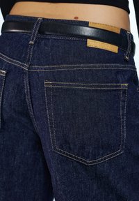 Tamno plave traperice s detaljnim prikazom stražnjih džepova, istaknute žutim šavovima i kožnim oznakom s logotipom marke na pojasu.