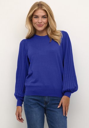 Blue knit sweater with ribbed puff sleeves and a round neckline. Paired with blue jeans, against a plain background.