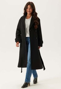 Cappotto in lana nera con design doppiopetto, ampi rever e tasche laterali, abbinato a jeans in denim blu e stivali neri con tacco.