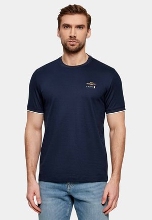 Uomo che indossa una maglietta a maniche corte blu navy con piccolo emblema dorato e testo "A.M.1923" sul petto sinistro, abbinata a jeans azzurri.