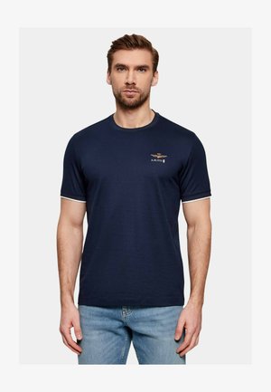 Uomo che indossa una maglietta a maniche corte blu navy con piccolo emblema dorato e testo "A.M.1923" sul petto sinistro, abbinata a jeans azzurri.