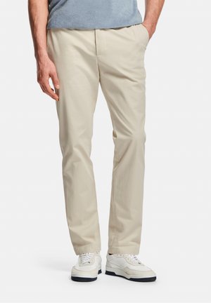 Homme portant un pantalon beige à jambes droites et des baskets blanches, debout avec une main dans la poche sur un fond blanc.