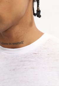 Camiseta branca com decote redondo, feita de um tecido leve e transparente. Tatuagem visível no pescoço com texto em caligrafia dizendo "acredita em ti mesmo."