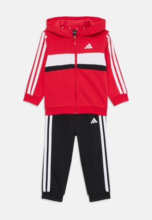 Raudona Adidas gobtuvu striukė su baltais ir juodais dryžiais, derinama su juodais treningais, turinčiais baltus dryžius ir logotipą. Sukurta aktyviam dėvėjimui.