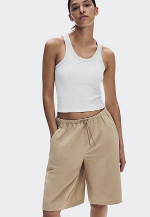 Young woman wearing a white sleeveless crop top and beige drawstring knee-length shorts, standing with hands in pockets.