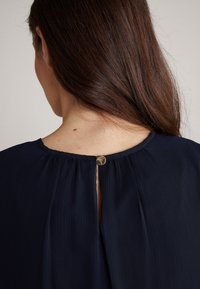 Navyblaues plissiertes Blusenshirt mit einem Schlüssellochausschnitt und einem goldenen Knopfakzent. Weicher Stoff mit einem gerafften Ausschnitt.