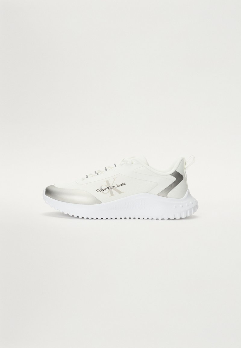 Zapatilla deportiva blanca con un suave acento plateado, suela texturizada y detalle del logo. Presenta un diseño de cordones y un talón acolchado para mayor soporte.