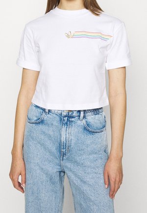 Personne portant un t-shirt court blanc avec un petit logo doré et des rayures horizontales arc-en-ciel, associé à un jean bleu clair taille haute.