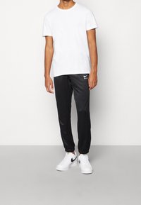 Man iförd en enkel vit t-shirt med kort ärm, svarta Nike Air joggers och vita Nike sneakers mot en enfärgad bakgrund.