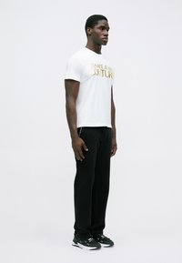 Muž v bílém tričku Versace Jeans Couture, černých kalhotách a černých teniskách, stojí před jednoduchým bílým pozadím.