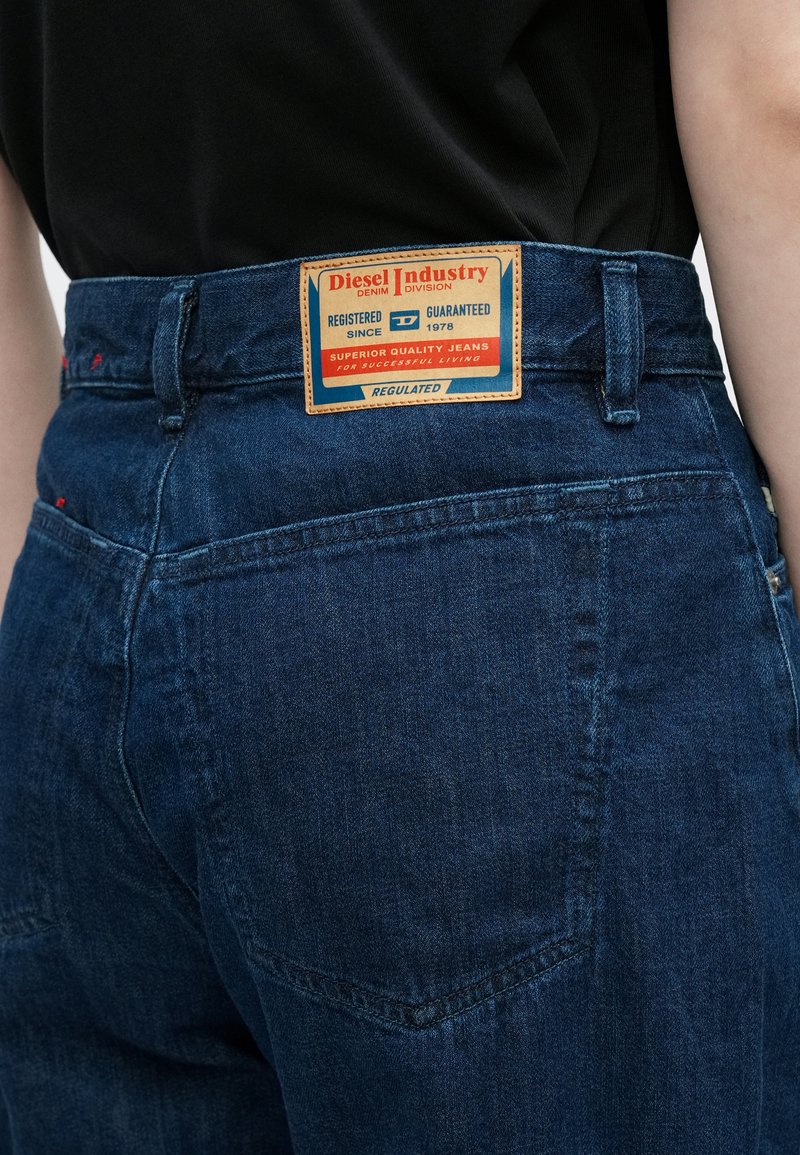Mørkeblå denimjeans med høj talje, der har en kvadratisk orange mærkat med mærket "Diesel Industry" og produktinformation.
