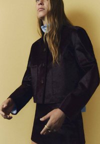 Giacca cropped borgogna con una texture liscia, caratterizzata da un colletto a punta, tasche frontali e chiusura con bottoni. Abbinata a una gonna coordinata.