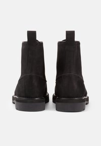 Botines de ante negro con una textura suave, punta redonda y suela de goma negra. Presentan una lengüeta en la parte trasera y detalles de costura sutiles.